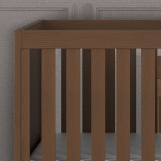 A close-up view of the Child Craft Kieran 3-in-1 Convertible Crib in Toasted Chestnut, featuring vertical slats and a gray mattress, set against a gray paneled wall.