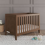 The Child Craft Kieran 3-in-1 Convertible Crib in Toasted Chestnut with a white mattress sits on a light rug in a gray-paneled room, next to a basket with a teddy bear, a toy stack, and an elephant plush on the floor.