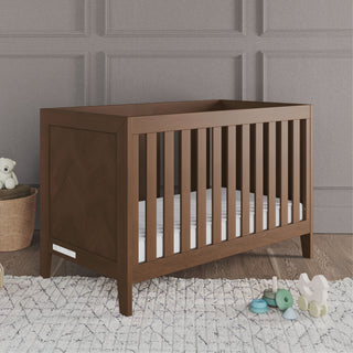 The Child Craft Kieran 3-in-1 Convertible Crib in Toasted Chestnut with a white mattress sits on a light rug in a gray-paneled room, next to a basket with a teddy bear, a toy stack, and an elephant plush on the floor.