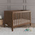 The Child Craft Kieran 3-in-1 Convertible Crib in Toasted Chestnut with a white mattress sits on a light rug in a gray-paneled room, next to a basket with a teddy bear, a toy stack, and an elephant plush on the floor.