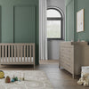 A modern nursery with green walls, a wooden crib, toy basket, teddy bear, and colorful toys on a patterned rug. A Child Craft F01792 Universal Dressing Kit in Crescent Grey adds style atop the dresser. Natural light streams in through a large window.