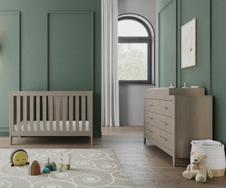 Modern nursery with green walls, a wooden crib, and the Child Craft Kieran 6-Drawer Double Dresser in Crescent Gray. Toys rest on a patterned rug, light pours through white curtains, and a teddy bear sits beside a white basket in the corner.