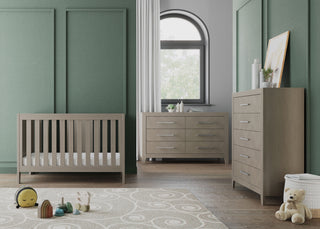 Modern nursery with sage green and light gray walls, a wooden crib, white curtains, a round swirl rug, plush toys on the floor, and the Child Craft Kieran 6-Drawer Double Dresser in Crescent Gray. Sunlight streams through a nearby window.