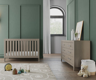 A modern nursery with green paneled walls features a wooden crib, the Child Craft Kieran 6-Drawer Double Dresser in Crescent Gray with decor and framed art, a toy bear by a basket, toys on a soft rug, and a large arched window with white curtains.