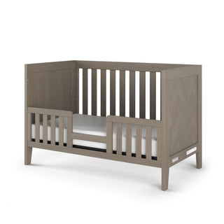 The Child Craft Kieran 3-in-1 Convertible Crib in Crescent Gray features slatted sides, a partial front rail, and a white mattress. Its modern, sturdy design transitions from crib to toddler bed for lasting versatility.