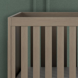 A close-up of the Kieran 3-in-1 Convertible Crib in Crescent Gray by Child Craft, featuring vertical wooden slats, set against a textured green wall.