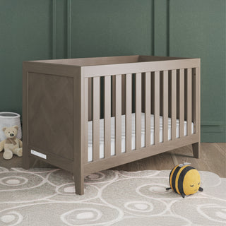 The Child Craft Kieran 3-in-1 Convertible Crib in Crescent Gray stands on a light patterned rug with white bedding. A teddy bear, white container, and plush bee toy add charm, while green walls with molding complete the modern nursery look.