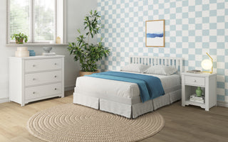 A bright bedroom features a bed, blue accents, white furniture including the Child Craft Harmony 3-Drawer Dresser with Changing Table Topper in matte white, a checkered blue and white wall, potted plant, round rug, natural light, and framed art above the bed.