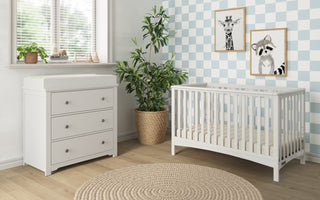 A modern nursery with a white crib, a Child Craft Harmony 3-Drawer Dresser with Changing Table Topper in matte white, a potted plant, animal artwork on blue-and-white checkered walls, bright sunlight, nursery storage, and a round beige rug on wood floors.