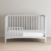 A Child Craft F09501 Toddler Guard Rail in matte white is attached to a white wooden toddler bed and placed against a plain light-colored wall on a wooden floor.