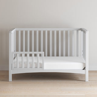 A Child Craft F09501 Toddler Guard Rail in matte white is attached to a white wooden toddler bed and placed against a plain light-colored wall on a wooden floor.