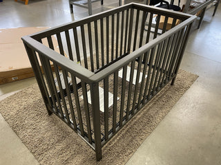 The Child Craft London 4-in-1 Convertible Crib in Dapper Gray, featuring baby-safe finishes and slatted sides, is displayed on a beige rug in a room with a concrete floor, with flat-pack furniture boxes and furnishings in the background.