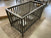 The Child Craft London 4-in-1 Convertible Crib in Dapper Gray, featuring baby-safe finishes and slatted sides, is displayed on a beige rug in a room with a concrete floor, with flat-pack furniture boxes and furnishings in the background.