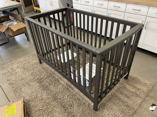 A Child Craft London 4-in-1 Convertible Crib in Dapper Gray sits on a beige rug in a room with white cabinets and boxes, appearing partially assembled.