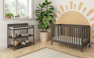 A nursery with a dark wood crib, Child Craft Flat Top Baby Changing Table in Dapper Gray, potted plant, sun mural, wooden floor, shelves for storage and toys, plus a window with blinds letting in natural light.