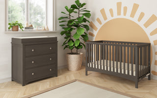 Modern nursery featuring the Child Craft London 4-in-1 Convertible Crib in Dapper Gray and matching changing table, with a potted plant, window blinds, and a sunburst wall design in warm tones finished with baby-safe paint.