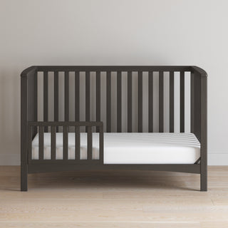 The Child Craft London 4-in-1 Convertible Crib in Dapper Gray features slatted sides, an open left section, baby-safe finishes, and a white mattress. It sits against a light wall on a light wood floor.