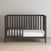 The Child Craft London 4-in-1 Convertible Crib in Dapper Gray features slatted sides, an open left section, baby-safe finishes, and a white mattress. It sits against a light wall on a light wood floor.