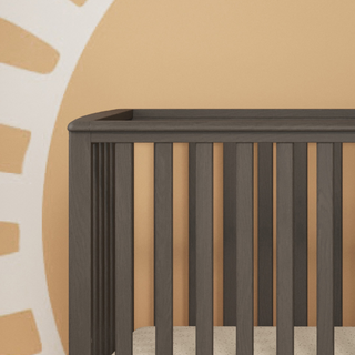 The Child Craft London 4-in-1 Convertible Crib in Dapper Gray, with baby-safe finishes and vertical slats, sits against a wall featuring a warm sunburst mural. A neutral mattress completes its modern look.