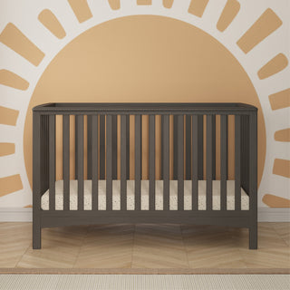 The Child Craft London 4-in-1 Convertible Crib in Dapper Gray stands sturdy by an orange sun mural. A patterned mattress and striped rug create a cozy feel on the wooden floor of this modern, baby-friendly nursery.