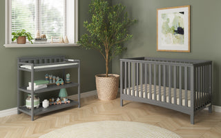 A modern nursery features sage green walls, a gray crib, and the Child Craft Flat Top Baby Changing Table in Cool Gray. Light wood herringbone floors, nursery storage, a potted plant, framed art, and baby items complete the decor.