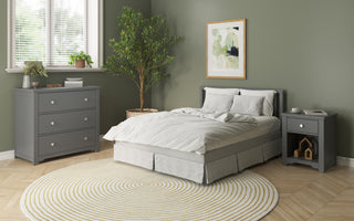 A cozy bedroom with green walls features a double bed with gray frame, white bedding, a Child Craft Harmony 3-Drawer Dresser with Changing Table Topper in Cool Gray, matching nightstand, nursery storage, plant, round rug, and abstract art above the bed. Sunlight streams in.