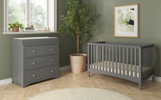 A nursery with sage green walls includes a gray crib, the Child Craft Harmony 3-Drawer Dresser with Changing Table Topper in Cool Gray, a kids dresser, a potted tree, window blinds, a round rug, and abstract art on the wall.
