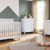 Modern nursery with a white crib, Child Craft F01792 Universal Changing Table Topper in Matte White on the dresser, and a changing table on wood flooring. Room features green walls, a patterned accent wall, beige curtains, wooden toys, and a round rug.