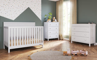 Modern nursery with a white crib, Child Craft F01792 Universal Changing Table Topper in Matte White on the dresser, and a changing table on wood flooring. Room features green walls, a patterned accent wall, beige curtains, wooden toys, and a round rug.