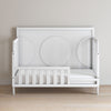 The Child Craft F09501 Toddler Guard Rail in matte white features sturdy wood construction, an open side with a simple slatted design, and circular back panel details, shown on a light wood floor against a plain beige wall.