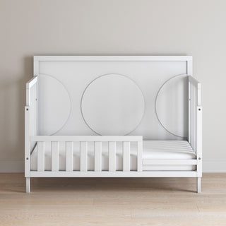 The Child Craft F09501 Toddler Guard Rail in matte white features sturdy wood construction, an open side with a simple slatted design, and circular back panel details, shown on a light wood floor against a plain beige wall.