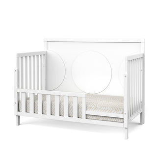 A Child Craft F09554 Toddler Guard Rail in matte white, shown with a modern crib featuring a patterned mattress and a headboard with two large circles.