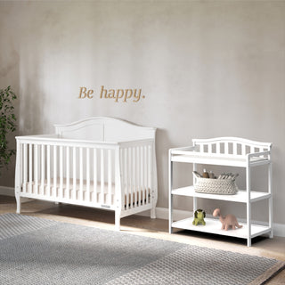 A white Child Craft Arch Top Baby Changing Table stands against a beige wall with “Be happy.” above. Baskets and toys fill the shelves, and a gray rug covers part of the wooden floor—perfect nursery furniture for your baby.