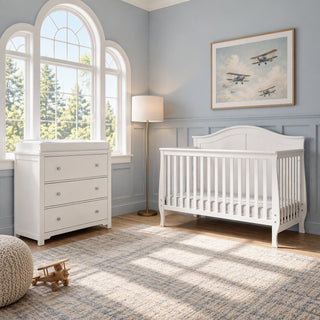 A white Child Craft Camden 2-Piece Nursery Set With Crib and Dresser featuring a convertible crib and a 3-drawer dresser with a changing table topper in a room with a window and framed airplane artwork.