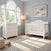 A white Child Craft Camden 2-Piece Nursery Set With Crib and Dresser featuring a convertible crib and a 3-drawer dresser with a changing table topper in a room with a window and framed airplane artwork.