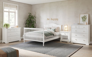 Bright bedroom with Child Craft Camden 4-in-1 Convertible Crib in matte white, matching nightstand and dressers, a large gray rug, potted plant, and "Be happy" wall art. Sunlight from the window creates a calm, cozy vibe.