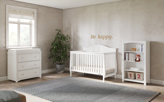 A nursery with beige walls, a white crib, Child Craft Harmony 3-Drawer Dresser with Changing Table Topper in matte white, a bookshelf with toys, gray rug, potted plant, window, and “Be happy.” above the crib in gold letters.
