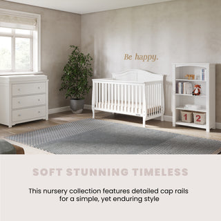 A modern nursery featuring the Child Craft Harmony 3-Drawer Dresser with Changing Table Topper in matte white, a white crib, spacious bookshelf, large potted plant, gray rug, and “Be happy.” on the wall, highlighting timeless, smart storage solutions.
