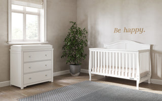 A bright nursery features a white crib, a Child Craft Harmony 3-Drawer Dresser with Changing Table Topper in Matte White, a potted plant, and a window. “Be happy.” is written on the beige wall, adding cheer and ample organized nursery storage.