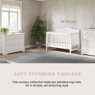 A bright nursery with white furniture, including the Child Craft Camden 4-in-1 Convertible Crib in Matte White, dresser, and chest. A plant is next to the crib, "Be happy." is on the wall, and there's a large window with a gray rug.