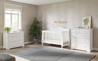 A bright nursery with beige walls, white furniture including the Child Craft Camden 4-in-1 Convertible Crib in matte white, two dressers, a potted plant, framed leaf print, and a rug. "Be happy." is written above the crib.