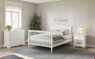 A bright, cozy bedroom with a white bed, nightstand, and dresser. A Child Craft crib with F06454 Full-Size Bed Rails in Matte White sits near a potted plant. “Be happy.” is written above the bed as soft natural light streams in.