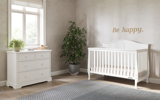 A bright nursery features a Child Craft Camden 4-in-1 Convertible Crib in matte white, a matching dresser with plants, a corner potted plant, and “Be happy.” on the wall. Sunlight streams through the window, lighting up the room.