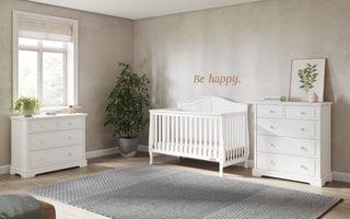 Bright nursery with a Child Craft Camden 4-in-1 Convertible Crib in Matte White, two white dressers, potted plant, framed leaf print, “Be happy.” wall text, and a large window letting in natural light.