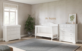 A bright nursery with white furniture, including the Child Craft Camden 4-in-1 Convertible Crib in Matte White, two dressers, a potted plant, a framed leaf print, and a rug. "Be happy." is displayed on the wall in warm-toned letters.