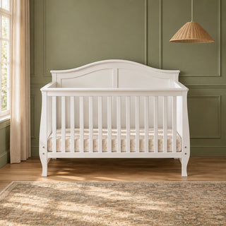 A matte white Child Craft Camden 4-in-1 Convertible Crib with a curved headboard and slatted sides, shown in a nursery.