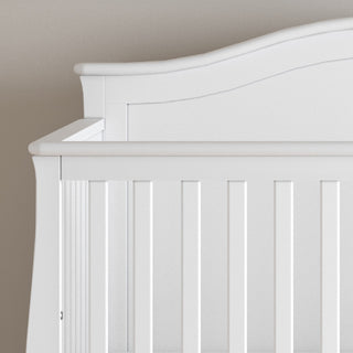 The partially visible Child Craft Camden 4-in-1 Convertible Crib in matte white, with vertical slats and a curved headboard, is set against a neutral wall for a timeless nursery look.