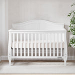 The Child Craft Camden 4-in-1 Convertible Crib in Matte White, featuring vertical slats and a curved headboard, is placed against a light wall on beige carpet, with a green potted plant partly visible on the right.