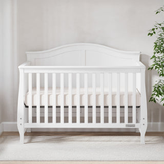 The Child Craft Camden 4-in-1 Convertible Crib in Matte White, featuring vertical slats and a curved headboard, is placed against a light wall on beige carpet, with a green potted plant partly visible on the right.