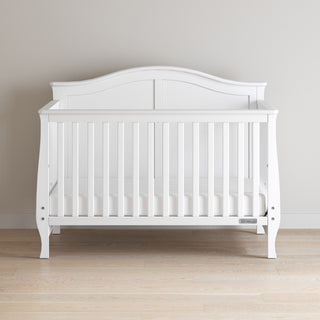 The Child Craft Camden 4-in-1 Convertible Crib in Matte White features a wooden frame with vertical slats and a curved headboard, shown on a light wood floor against a plain beige wall.
